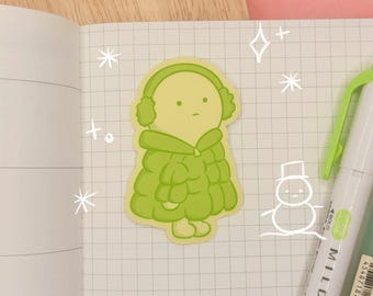 May include: A die-cut sticker featuring a light green cartoon character wearing earmuffs and a puffy jacket, placed on a grid-lined notebook page next to a white Mildliner highlighter with a small hand-drawn snowman doodle nearby.