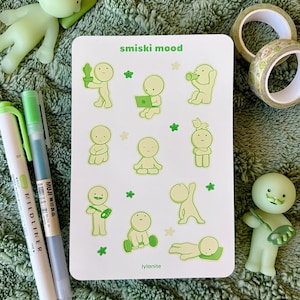 May include: A white sticker sheet with the text "smiski mood" and "lylanite" featuring green cartoon figures in various poses. Two green and white pens, two rolls of decorative tape, and two green figurines are also visible.