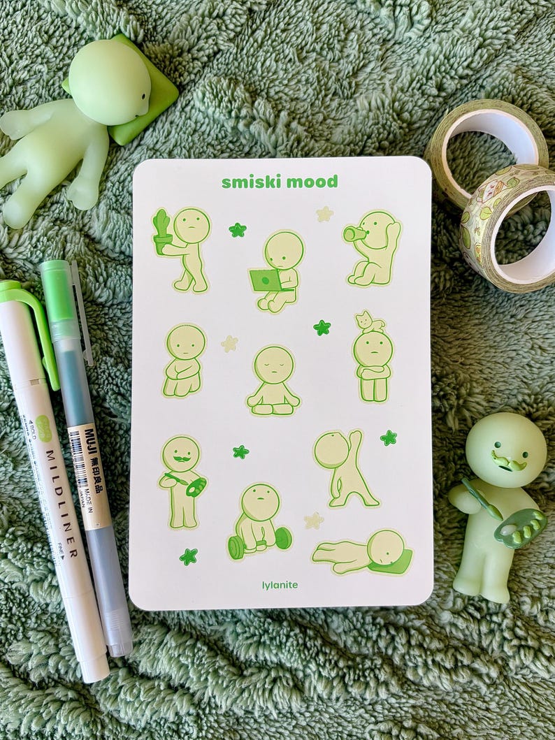 May include: A white sticker sheet with the text "smiski mood" and "lylanite" featuring green cartoon figures in various poses. Two green and white pens, two rolls of decorative tape, and two green figurines are also visible.