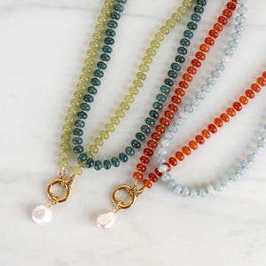 May include: Four beaded necklaces featuring small round stones in shades of light green, dark teal, orange, and pale blue. Each necklace includes a gold-toned circular clasp and a single white baroque pearl pendant hanging from the center.