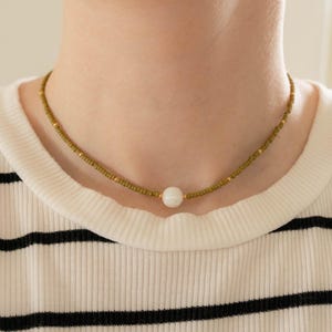 May include: A beaded choker necklace featuring small olive green seed beads strung along the length, accented with tiny gold-toned spacers and a single, smooth, round white bead positioned centrally as the focal point of the jewelry piece.