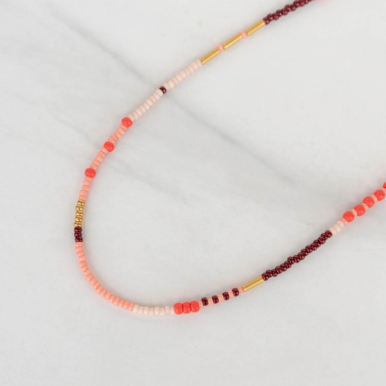 May include: A delicate beaded necklace featuring a mix of small, colorful beads. The necklace incorporates shades of pink, red, and gold, creating a vibrant and eye-catching design. The beads are strung together, forming a graceful curve. This necklace is a stylish accessory.