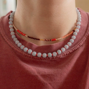 May include: Two necklaces are displayed. The lower necklace features large, light blue beads strung on red cord. The upper necklace has smaller, colorful beads in red, brown, and peach. The necklaces are worn on a person wearing a red t-shirt.