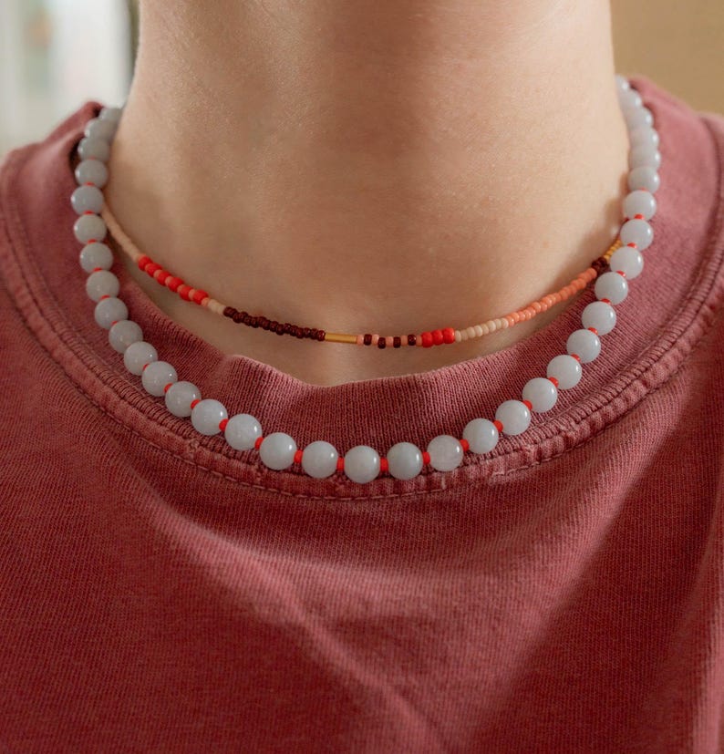 May include: Two necklaces are displayed. The lower necklace features large, light blue beads strung on red cord. The upper necklace has smaller, colorful beads in red, brown, and peach. The necklaces are worn on a person wearing a red t-shirt.
