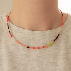 May include: A beaded necklace featuring a mix of small round beads in shades of coral, red, dark maroon, white, and lime green, interspersed with thin gold-toned cylindrical spacers, arranged in a continuous circular pattern around the neck.
