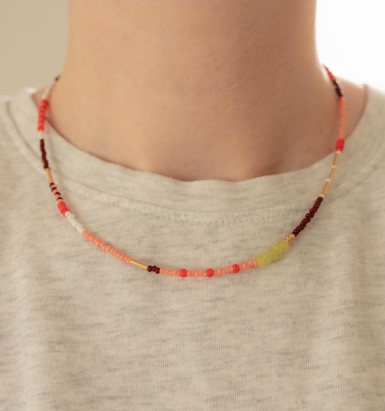 May include: A beaded necklace featuring a mix of small round beads in shades of coral, red, dark maroon, white, and lime green, interspersed with thin gold-toned cylindrical spacers, arranged in a continuous circular pattern around the neck.