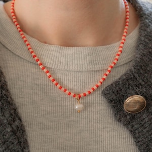 May include: A beaded necklace featuring alternating small cylindrical beads in bright orange and pale peach colors. The necklace includes a single white, oval-shaped pearl pendant suspended from a small gold-toned metal ring at the center.