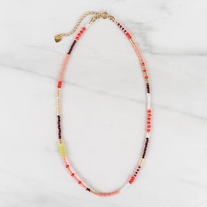 May include: A beaded necklace featuring a sequence of small round beads in shades of coral, dark red, white, and yellow, interspersed with thin gold-toned metal cylindrical spacers. The necklace includes a gold-toned metal chain extender and clasp.