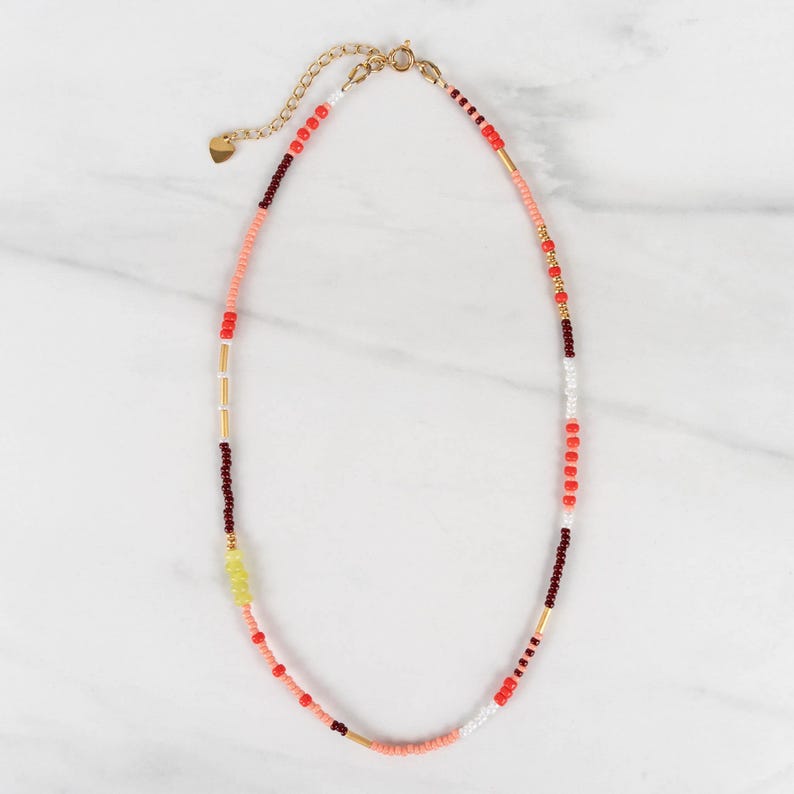 May include: A beaded necklace featuring a sequence of small round beads in shades of coral, dark red, white, and yellow, interspersed with thin gold-toned metal cylindrical spacers. The necklace includes a gold-toned metal chain extender and clasp.