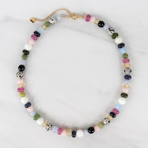 May include: A beaded necklace featuring a variety of colorful, smooth, and speckled round beads in shades of pink, blue, green, black, white, and tan. The necklace includes a gold-toned metal clasp and an adjustable chain with a small heart charm.