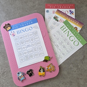 May include: A pink pin trading Bingo board with attached pins, and three Bingo cards. The cards are labeled "Magic Kingdom", "Hollywood Studios", "Animal Kingdom", and "Epcot". The pins include cartoon characters and a hat.