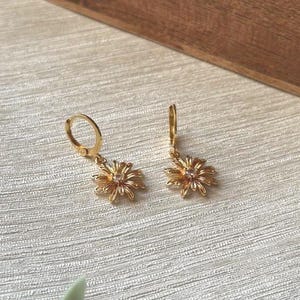 Puede incluir: Un par de pendientes de aro dorados con un colgante de flor. El colgante de flor tiene un pequeño strass transparente en el centro.