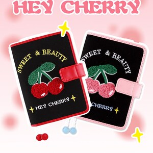 Könnte beinhalten: Zwei schwarze Stoff-Notizbücher mit roten und rosafarbenen Lederelementen. Beide Notizbücher haben gestickte Kirschen und den Text "Sweet & Beauty" und "Hey Cherry".