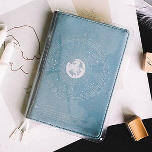 May include: A light blue notebook with a clear plastic cover. The cover features a white design of a circle with stars and a globe inside.