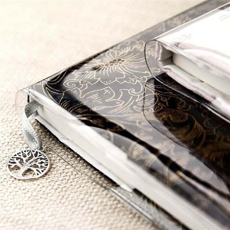 May include: A black and gold patterned journal with a clear plastic cover and a silver tree of life charm attached to a silver ribbon bookmark.