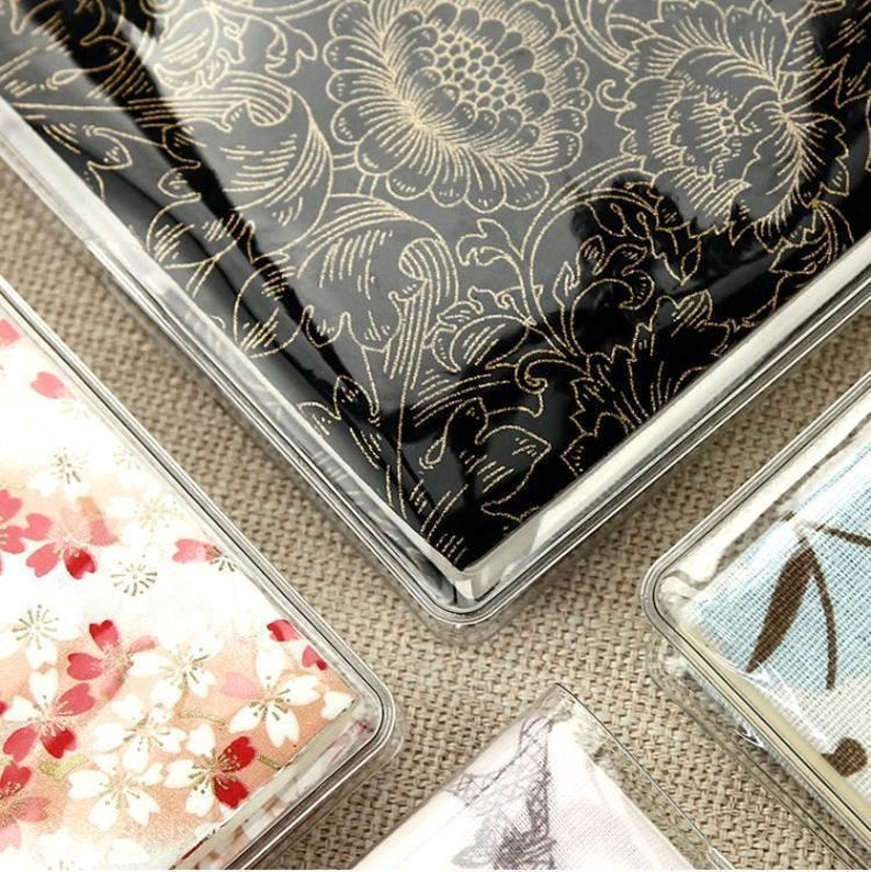 May include: Four clear plastic cases with different patterned fabric inserts. The cases are square and have a silver trim. The patterns include a black floral design, a white floral design, a blue and white floral design, and a white and gray butterfly design.