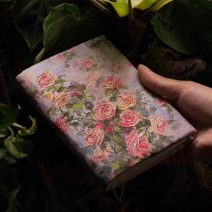 May include: A small, hand-painted notebook with a floral design featuring pink and yellow roses on a light purple background. The notebook is held in a hand.
