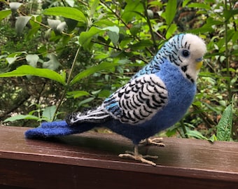 Puede incluir: Un periquito azul y blanco de fieltro de aguja posado sobre una superficie de madera. El periquito tiene un cuerpo azul claro con marcas blancas y negras en sus alas.