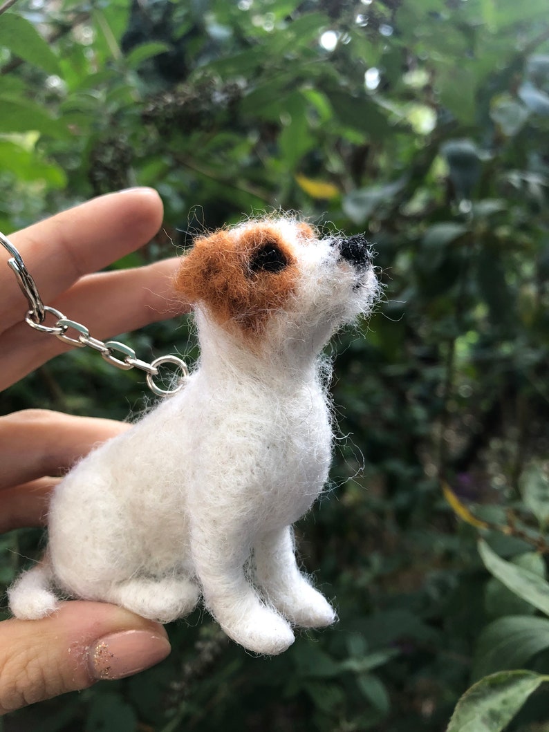 May include: A white and brown felt dog keychain with black eyes. The dog is sitting up and looking to the right. The keychain is attached to a silver chain.