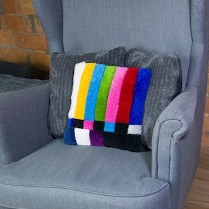May include: A grey armchair with a high back and wing arms. The chair has a colourful pillow with a television test pattern design on it.