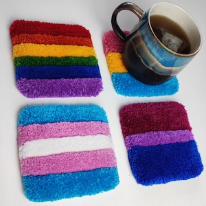 Puede incluir: Cuatro posavasos cuadrados de crochet con rayas de diferentes colores. Los posavasos son de los colores de las banderas del arcoíris, transgénero y bisexual. Una taza azul con té está sobre uno de los posavasos.