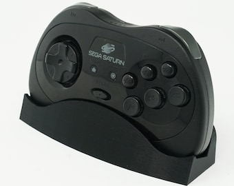Puede incluir: Un mando negro de Sega Saturn con una cruceta, botones y el logotipo de Sega Saturn. El mando se muestra sobre un soporte negro sobre un fondo blanco. El texto "SEGA SATURN" es visible en el mando.