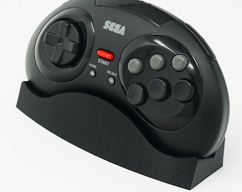 Puede incluir: Un mando negro de Sega Genesis con un botón de inicio rojo y una base negra. El mando tiene un pad direccional, seis botones y el texto "SEGA" impreso en la parte superior.