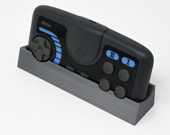 Puede incluir: Un mando 8BitDo negro y gris con un panel de botones azul y gris, un pad direccional gris y dos botones grises etiquetados como "I" y "II". El mando está en un soporte gris.