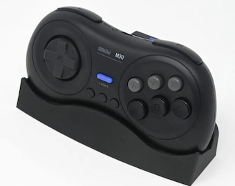 Puede incluir: Un mando negro de estilo Sega Genesis con un botón de inicio azul y el texto "8BitDo M30" en la parte delantera. El mando está colocado en un soporte negro.