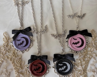 May include: Four silver-toned necklaces featuring circular pendants with wax seal-style designs of a crescent moon and bats. Each pendant is accented with a black ribbon bow and small bat charms on the chain. The pendants come in lavender, red, black, and pink.