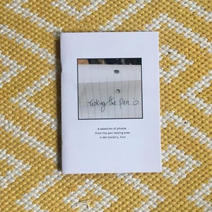 May include: A white booklet with the text "Testing the pen" and a photo of a piece of paper with two holes punched in it. The text below the photo reads "A selection of photos from the pen-testing area in WH Smith's, York."
