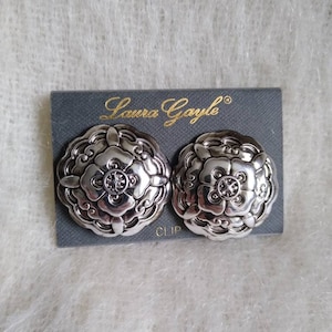 May include: A pair of silver-toned clip-on earrings with a floral design. The earrings are round with detailed petal-like edges and a central design. The earrings are on a gray card with the text "Laura Gayle" and "CLIP".