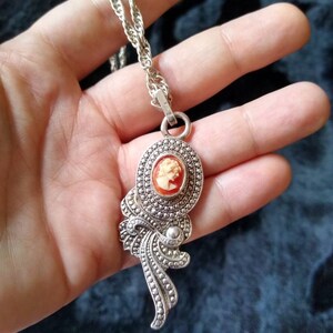 May include: A silver chain necklace with a cameo pendant. The pendant features a detailed design with a cameo of a woman's face.