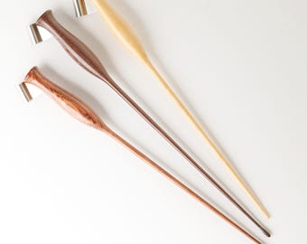 May include: Three wooden drop spindles with tapered handles and metal hooks at the top. The spindles are arranged diagonally, featuring light, medium, and dark brown wood grains with smooth, polished finishes and slender, pointed tips.