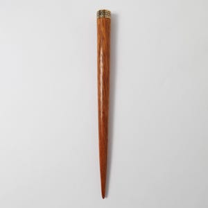 Könnte beinhalten: Ein langer, sich verjüngender Haarstab aus Holz mit rotbraunem Finish. Der obere Teil hat ein dekoratives goldfarbenes Band mit einem sich wiederholenden geometrischen Muster. Der Stab ist unten spitz und zum Haare stylen gedacht.
