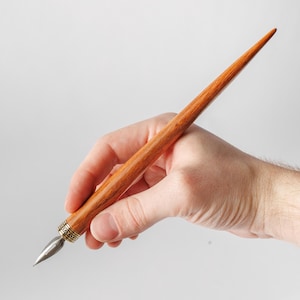Puede incluir: Una pluma de caligrafía con un mango de madera liso y cónico de veta marrón cálida. La pluma está equipada con una punta de metal plateado y una banda decorativa de metal dorado grabado que une la punta al cuerpo de madera.