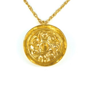 May include: A gold-toned pendant necklace featuring a circular pendant with an intricate, embossed design. The pendant has a central raised circle surrounded by swirling patterns. The necklace chain is also gold-toned and appears to be a rope style.