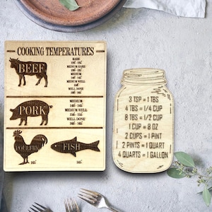 May include: Wooden kitchen sign with cooking temperatures for beef, pork, poultry, and fish. A separate wooden sign with a mason jar graphic shows common cooking measurements in US customary units.