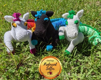 May include: Three crocheted stuffed animals, a white unicorn, a black horse, and a green and blue dragon, are lying on a bed of green grass. The unicorn has a pink mane and tail, the horse has a blue mane and tail, and the dragon has green scales and a blue belly. The animals are all looking at the camera.