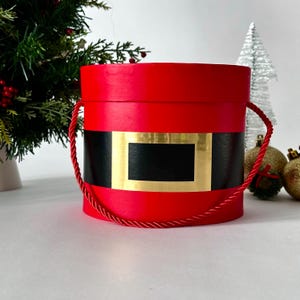May include: A round red gift box with a black band and gold buckle design, resembling Santa's belt. It has a red rope handle and is decorated with Christmas ornaments and a small Christmas tree.