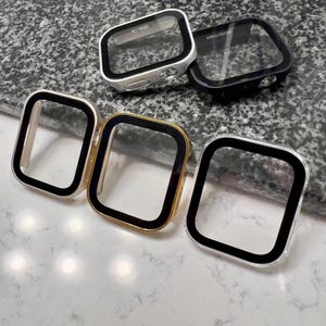 May include: A collection of Apple Watch screen protectors in various colors. The protectors have a clear screen with a black border and are available in silver, black, and gold. The protectors are square-shaped and designed to fit the Apple Watch.