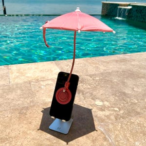 May include: A black smartphone mounted on a silver metal stand, featuring a pink sun umbrella attached to the back of the device via a circular red magnetic mount. The umbrella has a flexible stem and white tips on the ribs, providing shade.