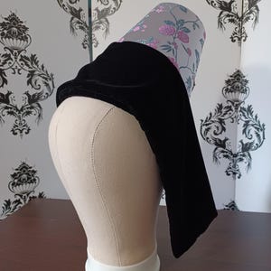 May include: A black velvet hat with a floral patterned brim. The hat is displayed on a mannequin head. The brim features a gray background with pink and purple floral designs. The hat's design is a combination of classic and modern styles.