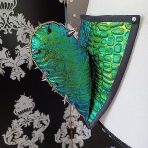 May include: A close-up of a green and gold textured fabric with silver spikes. The fabric has a scale-like pattern and is trimmed with black fabric. The design is attached to a white surface.
