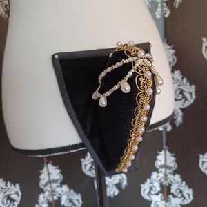 May include: A black velvet headband with gold trim and faux pearls. The headband is adorned with multiple strands of small pearls and larger teardrop-shaped pearls. The gold trim has an ornate, filigree design. The headband is displayed on a mannequin.
