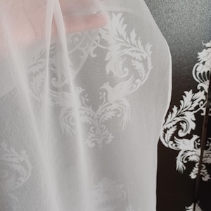 Puede incluir: Tela blanca transparente con un patrón floral y de pájaros repetido. La tela tiene una caída suave y permite el paso de la luz. El diseño es de estilo clásico y elegante, adecuado para diversos usos decorativos.