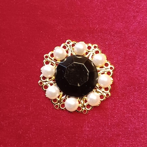 May include: A gold-toned brooch with a central black gemstone surrounded by white pearls. The brooch has an ornate, openwork design and is set against a red background.