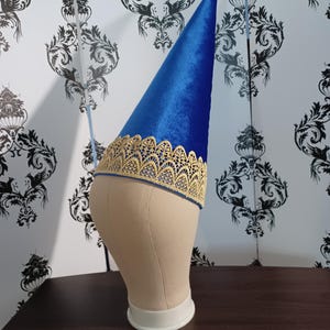 May include: A blue velvet wizard hat with a gold lace trim, displayed on a mannequin head. The hat has a tall, pointed crown and a decorative band around the base. The background features a black and white floral wallpaper.