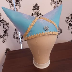 May include: A light blue, two-pointed headpiece featuring a shimmering fabric finish. The hat is adorned with intricate gold-colored lace trim along the edges and a single, circular, light blue rhinestone brooch centered on the front panel.