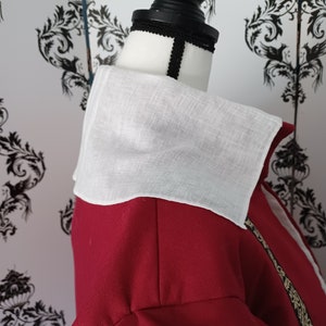 Puede incluir: Una camisa roja con cuello, un cuello de lino blanco y ribetes dorados. La camisa está en un maniquí blanco.